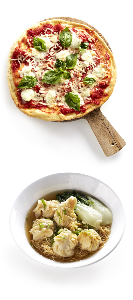 A pizza on a white plate next to a bowl of soup placed on a river rock.
